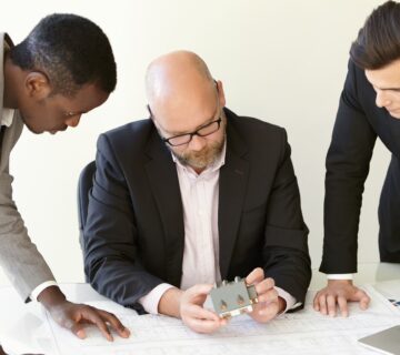 Shot working process construction design bureau three men office clothes examining new perspective project main design engineer sitting table his coworkers standing aside him 1 360x320